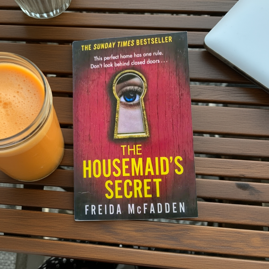Book cover of 'The Housemaid's Secret' by Freida McFadden on a table with a juice