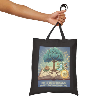 Black tote bag with a colorful design of an open book with a tree and characters, held by a hand against a white background.