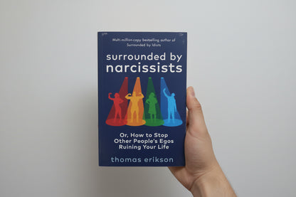 Book titled 'Surrounded by Narcissists' held by a hand against a blue background